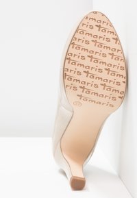Chaussure à talon haut beige à bout pointu, présentée de biais pour montrer la semelle avec le logo "tamaris" répété et la taille 37 près de l'arrière du talon.