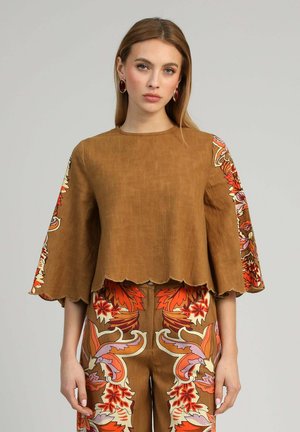 Femme portant un haut marron avec des manches larges à motif floral et un pantalon assorti à motif floral sur un fond gris uni.