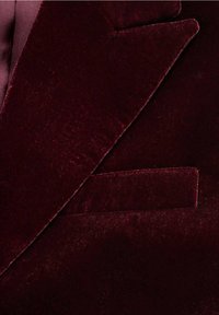 Gros plan du revers et de la poche poitrine d'un blazer en velours bordeaux foncé, montrant une texture douce et des détails de couture fins.