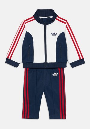 Tuta sportiva navy e bianco con strisce rosse, giacca con zip e colletto a coste, e pantaloni abbinati con logo sul lato.