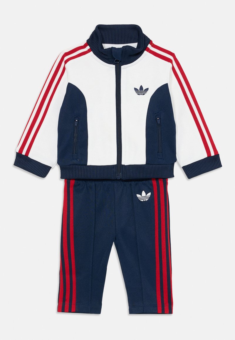 Survêtement de sport bleu marine et blanc avec des rayures rouges, blouson zippé avec col côtelé et pantalon assorti avec logo sur le côté.