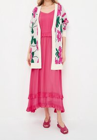 Lange roze jurk met ruches aan de zoom, gelaagd onder een witte cardigan met grote roze bloemenborduursels. Draagt roze open-toe schoenen.