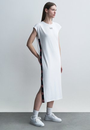 Femme portant une robe midi sans manches blanche avec des bandes latérales noires et des logos EA7, debout avec une main derrière le dos, baskets blanches et chaussettes.