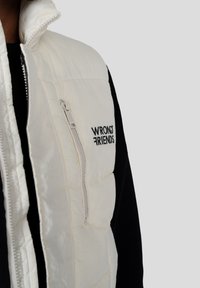 Nahaufnahme einer Person, die eine weiße Steppweste mit dem Text "WRONG FRIENDS" und einer vertikalen Reißverschlusstasche auf der Brust trägt.
