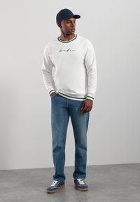 Vit sweatshirt med gröna detaljer och broderad text, kombinerad med blå jeans och blå sneakers. Man står med händerna i fickorna.