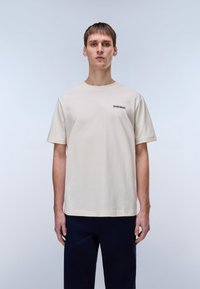 Φωτεινό μπεζ βαμβακερό μπλουζάκι με στρογγυλή λαιμόκοψη, κοντά μανίκια και ένα μικρό μαύρο γράφημα στο στήθος. Απλός, ευθύγραμμος σχεδιασμός.