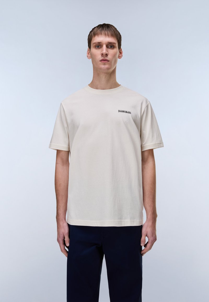 Φωτεινό μπεζ βαμβακερό μπλουζάκι με στρογγυλή λαιμόκοψη, κοντά μανίκια και ένα μικρό μαύρο γράφημα στο στήθος. Απλός, ευθύγραμμος σχεδιασμός.