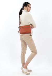 Leather crossbody bag in burnt orange with a zippered pocket and adjustable strap. Model wears cream sweater and beige pants, paired with sneakers.