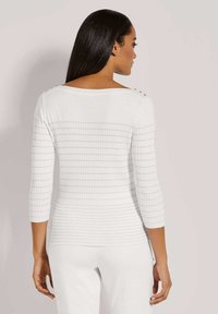 Weißer gerippter Pullover mit Dreiviertelärmeln, der eine figurbetonte Passform hat und goldene Knopf-Details an den Schultern aufweist. Der Stoff wirkt glatt.