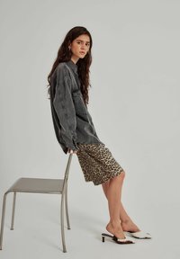 Übergroßes graues Denim-Hemd, Bleistiftrock in Leopardenmuster und weiße Blockabsätze. Texturen umfassen Denim und glattes Gewebe. Einfacher Hintergrund.
