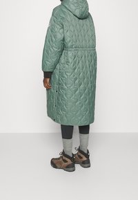 Persona che indossa un lungo cappotto a vento verde con cappuccio, leggings neri, calzini grigi e stivali da trekking marroni, in piedi contro uno sfondo semplice.