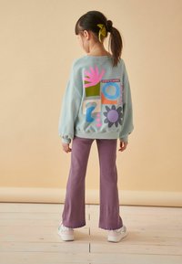Sweatshirt bleu clair avec des motifs géométriques colorés dans le dos, associé à un pantalon évasé violet et des baskets blanches avec des accents pastel.