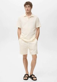 Polo a maniche corte e pantaloni coordinati in tessuto testurizzato off-white; presenta un colletto, vestibilità comoda e vita elasticizzata. Indossato con sandali neri.