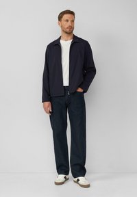 Marineblauwe jas met kraag, wit t-shirt, donkere spijkerbroek en witte sneakers met bruine accenten. Model staat tegen een effen achtergrond.