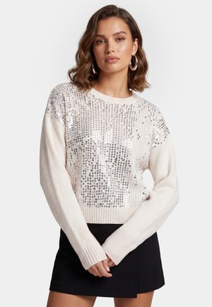 Vrouw met bruin golvend haar draagt grote oorringen, een witte trui met zilveren pailletten en een zwarte rok, staat met de handen gevouwen.