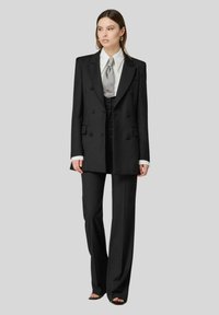 Blazer nero doppiopetto con rever a punta, abbinato a pantaloni neri a vita alta. Camicia bianca con cravatta grigia. Texture del tessuto liscia.