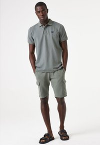 Groene, gestructureerde polo met korte mouwen en een logo op de borst, gecombineerd met bijpassende cargoshorts en zwarte sandalen. Eenvoudig, functioneel ontwerp.