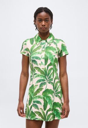 Moteris, vilkinti trumpomis rankovėmis su apykakle suknele, puošta dideliais žaliais tropinių lapų raštais ant šviesaus fono, stovinti priešais paprastą foną.