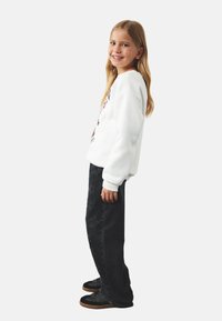 Hvid sweatshirt med grafisk design, sorte jeans med blomstermønster og sorte sneakers med gummisål. Barnet står sidelæns og smiler.