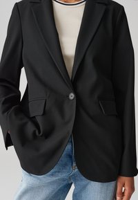 Zwarte op maat gemaakte blazer met een enkele knoopsluiting, twee voorzakken en een soepele stof. Gecombineerd met een ribbelbeige shirt en spijkerbroek.