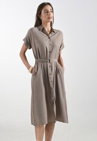 Modella che indossa un vestito button-up a maniche corte di colore marrone chiaro con una cintura in vita, in piedi con le mani nelle tasche su uno sfondo bianco.