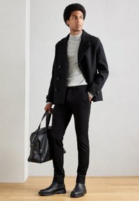 Abrigo negro de doble botonadura sobre un cuello de tortuga gris, pantalones negros ajustados y botas negras de tobillo, sosteniendo un bolso de cuero negro. Fondo de suelo de madera.
