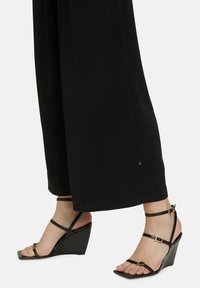 Zwarte wedge-sandalen hebben een glanzende afwerking, verstelbare enkelriemen en een vierkante open teenontwerp. Gecombineerd met zwarte wijde broek.