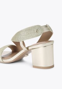 Sandales à talons dorés avec une sangle tissée et des accents métalliques. Design à talon carré avec une texture lisse et un bout ouvert.
