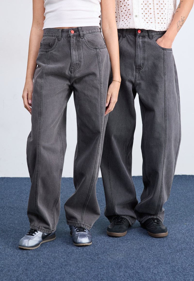 Jeans en denim gris clair à jambes larges avec des coutures prononcées, des poches avant et des accents de boutons rouges. Deux modèles portent des chaussures contrastantes.