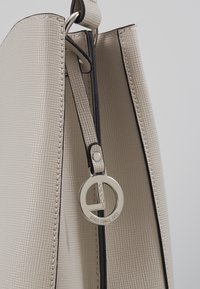 Sac à main structuré beige avec une surface texturée, des bords noirs, une forme élégante et une étiquette en métal argenté portant le logo de la marque.