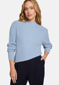 Pull en maille côtelée bleu clair avec un col montant et des manches bouffantes. Porté avec un pantalon sombre ajusté, affichant une coupe décontractée.