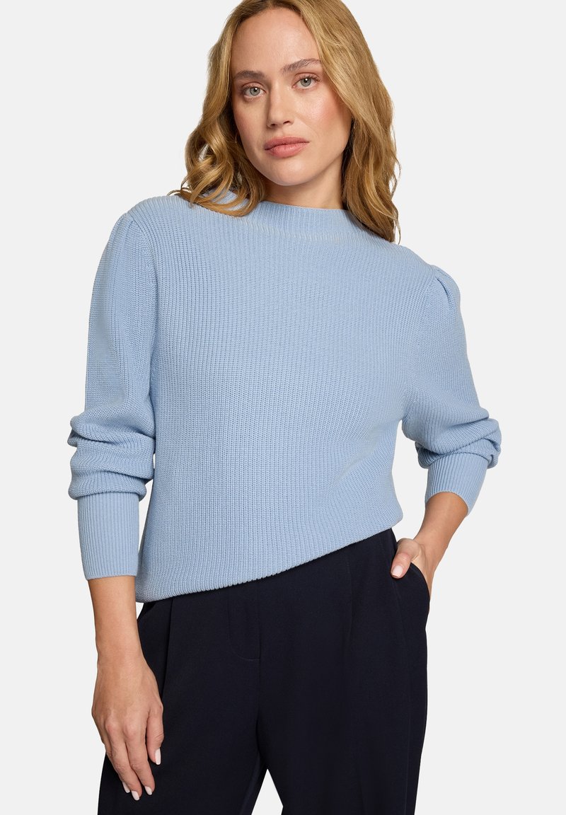 Pull en maille côtelée bleu clair avec un col montant et des manches bouffantes. Porté avec un pantalon sombre ajusté, affichant une coupe décontractée.