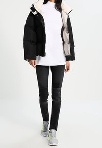 Chaquetón acolchado negro y beige con un diseño corto, camiseta blanca por debajo, jeans negros ajustados y zapatillas grises con acentos en neón.