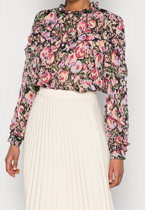 Blouse fleurie transparente à manches longues avec volants, associée à une jupe blanche plissée taille haute portée par une personne debout.
