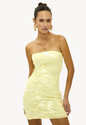 Femme avec les cheveux en chignon portant de grandes boucles d'oreilles en forme de fleur en or et une robe sans manches de couleur jaune clair avec de la broderie florale, posant les mains sur les hanches.