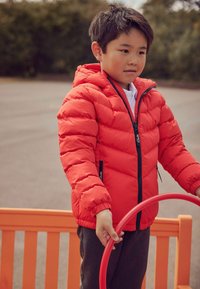 Czerwona kurtka puchowa z kapturem, z czarnym zamkiem błyskawicznym i elastycznymi mankietami, noszona na białej koszuli. Szare spodnie i czerwony pierścień w rękach.