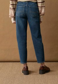 Donkerblauwe denim jeans met rechte pijpen, zichtbare stiksels en twee achterzakken, gecombineerd met bruine platform schoenen tegen een neutrale achtergrond.