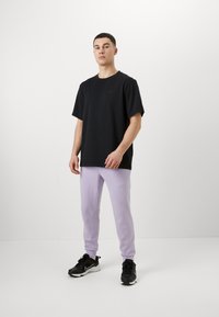 Black short-sleeve t-shirt, loose fit, worn with lilac sweatpants. Model stands on a white background, wearing black athletic shoes.