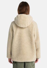 Persona che indossa un giubbotto con cappuccio, di colore crema e molto morbido, visto da dietro, abbinato a pantaloni cargo di colore verde oliva.