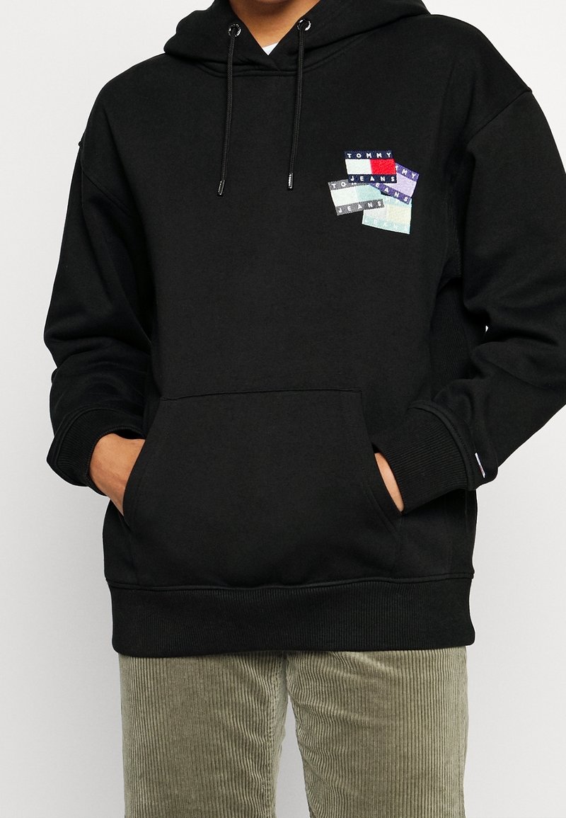 Sudadera con capucha negra y bolsillo frontal, con un parche de logo multicolor en el pecho. Puños y dobladillo de canalé, textura de tela suave.