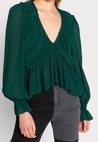 Blusa verde, con texture, scollatura a V profonda, dettagli arricciati sulle spalle e maniche lunghe a campana, abbinata a jeans neri a vita alta.