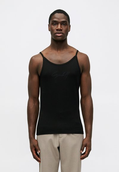 Must värviline sobiv tank top õhukeste rihmadega seisev mees, kes kannab beeže pükse, ühtlasel valgel taustal.