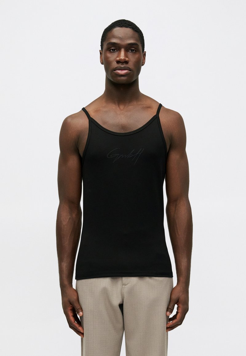 Canotta nera attillata con spalline sottili su un uomo in piedi che indossa pantaloni beige, di fronte a uno sfondo bianco semplice.