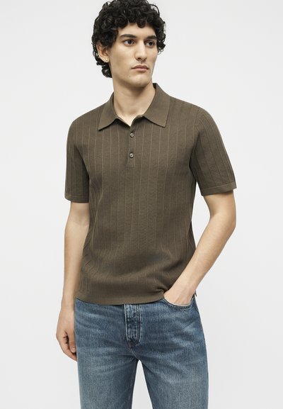 Νέος άνδρας με σγουρά μαλλιά που φορά καφέ κοντομάνικο πόλο με υφασμάτινες ρίγες και μπλε τζιν, ένα χέρι στην τσέπη, ουδέτερο φόντο.