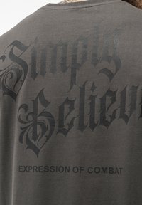 Pilkšvas medvilnės marškinėliai su dideliu tamsiu tekstu, "Tiesiog tikėk" ir "KOVOS IŠRAIŠKA", su puošnia lettering ir glotnia tekstūra.
