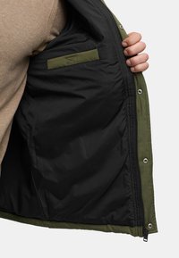 Olivgrüne Jacke mit schwarzem Futter, die einen Reißverschluss und eine Innentasche hat. Das Material wirkt glatt mit einer strukturierten Oberfläche.