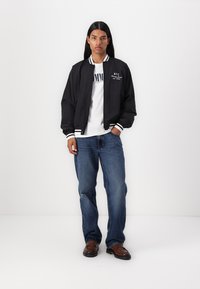 Sort bomberjakke med hvide stribede manchetter, båret over en hvid grafisk t-shirt og blå denim jeans. Modellen står foran en neutral baggrund.