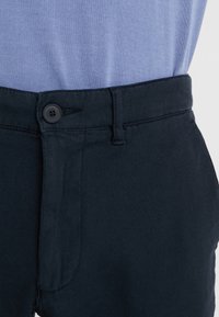 Zblízka černý zapínací pás na tmavých kalhotách nosených s vyčesanou světle modrou košilí.