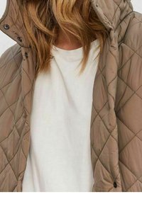 Chaqueta beige acolchada con capucha, con botones de presión y una textura suave. Llevada sobre una camisa blanca, destacando un diseño en capas.