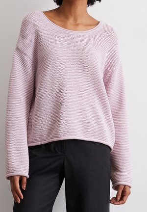 Personne portant un pull en tricot rose clair à manches longues avec un pantalon noir, debout devant un fond clair uni.
